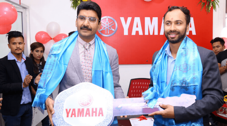 Two men standing in a yamaha showroom during a special event or ceremony, one holding a memento, both adorned with ceremonial scarves, with onlookers in the background.