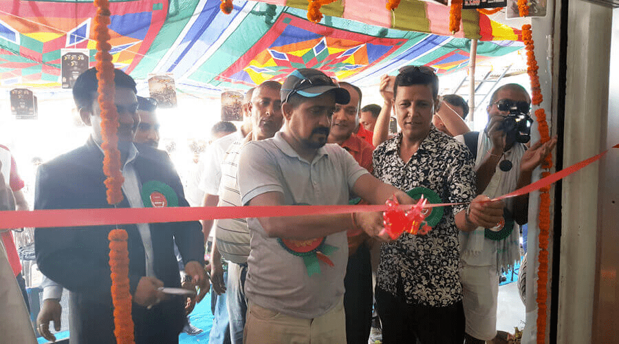 Two men hold a ceremonial ribbon, one of them ready to cut it with scissors, marking the grand opening of an event or establishment, as attendees watch and a photographer captures the moment.