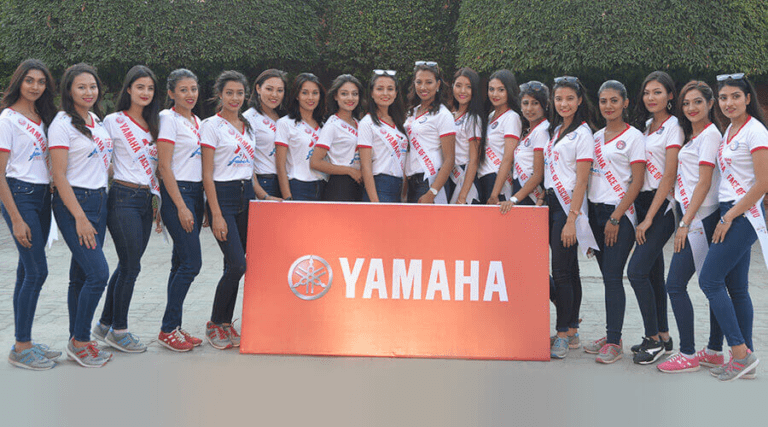 A group of smiling young women standing in a line, each wearing a sash and similar outfits, posing for a photo behind a large sign with the yamaha logo.