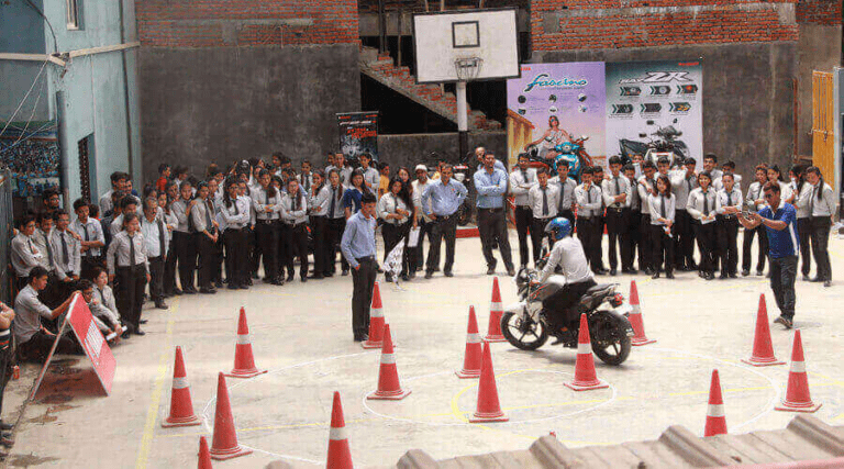 A motorcycle safety demonstration taking place in front of an attentive group of students and faculty, as a rider weaves through a course of orange traffic cones.